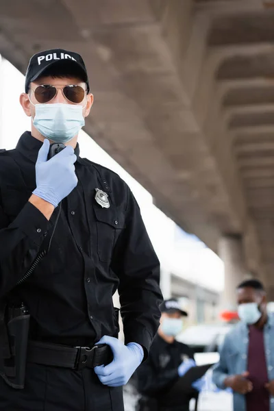 Oficial de policía con máscara médica y guantes de látex usando walkie talkie cerca de colega y hombre afroamericano sobre fondo borroso en la calle urbana - foto de stock