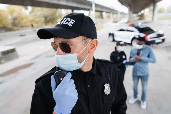 Policía en guante de látex y máscara médica usando walkie talkie cerca de colega y víctima afroamericana sobre fondo borroso en la calle urbana - foto de stock