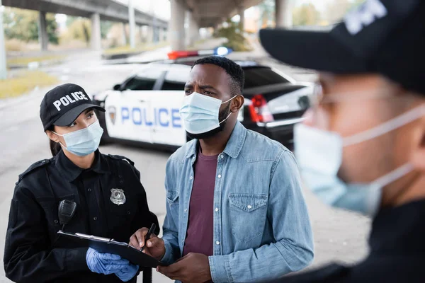 Víctima afroamericana en máscara médica sosteniendo pluma cerca de oficial de policía con portapapeles y policía en primer plano borroso - foto de stock