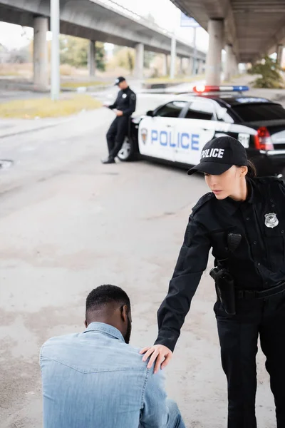 Police officer calming african american man near colleague and car on blurred background on urban street — Stock Photo