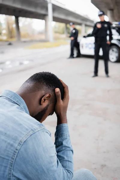 Triste víctima afroamericana cerca de oficiales de policía y coche sobre fondo borroso al aire libre - foto de stock
