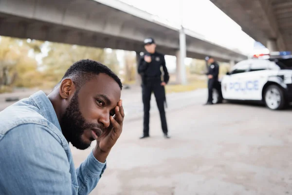Perturbada víctima afroamericana mirando hacia otro lado cerca de los oficiales de policía y el coche sobre fondo borroso en la calle urbana - foto de stock