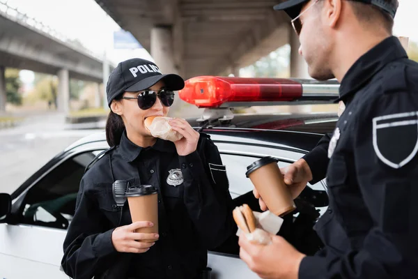 Mujer policía comiendo hamburguesa y sosteniendo café para ir cerca de colega y coche de policía en un fondo borroso - foto de stock