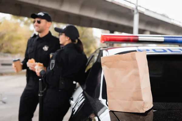 Паперовий пакет на багажнику автомобіля біля поліцейських з гамбургерами та кавою, щоб піти на розмитому тлі на міській вулиці — стокове фото