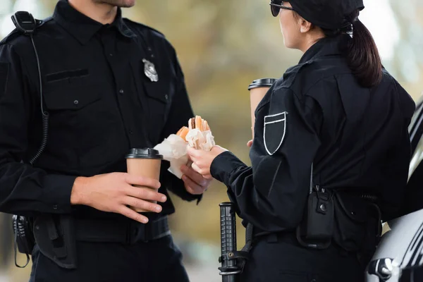 Женщина-полицейский в форме держит бургер и кофе на вынос рядом с коллегой и машиной на размытом переднем плане — стоковое фото