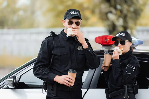 Policías en gafas de sol comiendo hamburguesas y sosteniendo café para llevar cerca del coche al aire libre - foto de stock