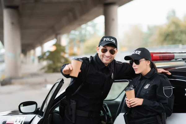 Cheerful police officer pointing with hand near colleague holding coffee to go and car outdoors — Stock Photo