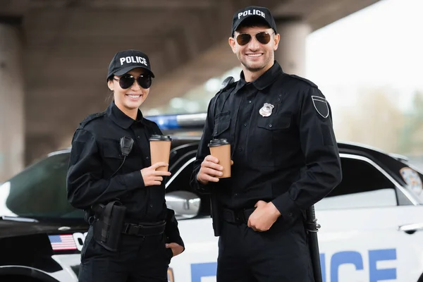Smiling police officers in sunglasses holding takeaway coffee near car on blurred background outdoors — Stock Photo