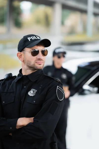 Policeman in sunglasses standing with crossed arms near colleague and auto on blurred background — Stock Photo