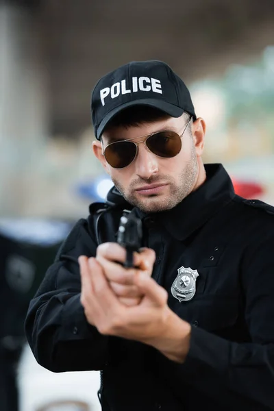 Police officer in uniform and sunglasses holding gun on blurred foreground outdoors — Stock Photo