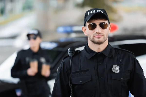 Офицер полиции в солнечных очках рядом с коллегой и автомобилем на размытом фоне на улице — стоковое фото