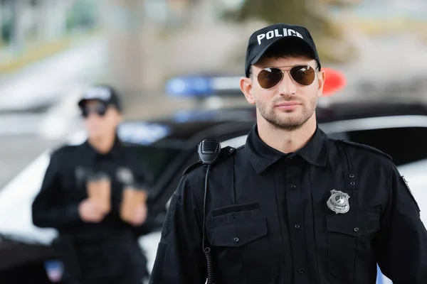 Oficial de policía en gafas de sol cerca de colega y coche sobre fondo borroso al aire libre - foto de stock