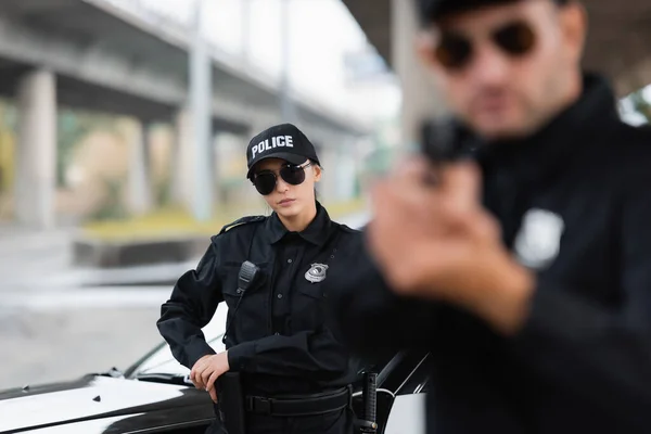 Mujer policía en gafas de sol tomando pistola cerca de coche y colega en primer plano borrosa al aire libre - foto de stock