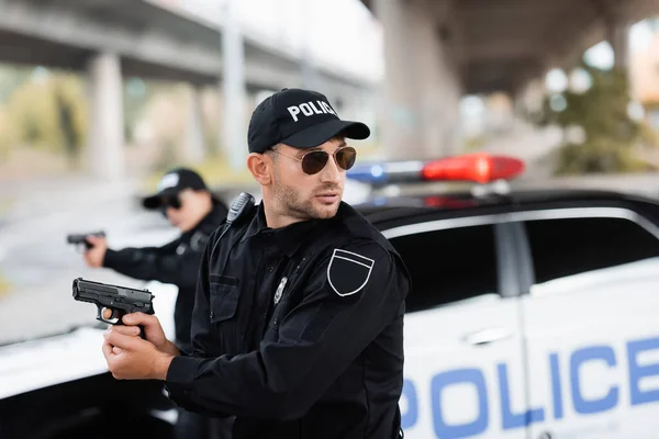 Policeman in sunglasses holding gun and looking away near colleague and car on blurred background — Stock Photo