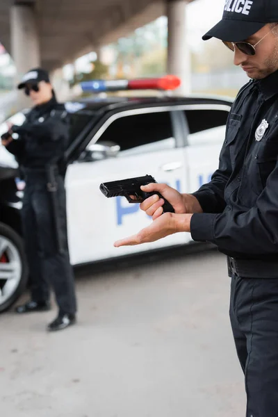 Офицер полиции заряжает пистолет рядом с коллегой и машиной на размытом фоне на городской улице — стоковое фото