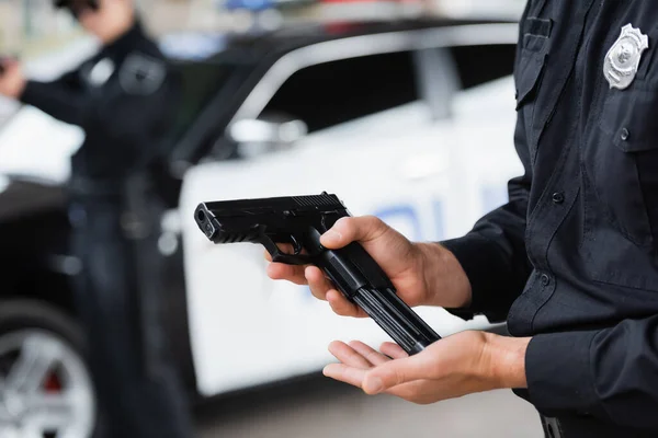 Vista ritagliata del poliziotto pistola di carico vicino collega e auto della polizia su sfondo sfocato — Foto stock