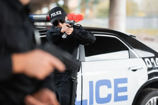 Policewoman in sunglasses holding gun near colleague on blurred foreground and car outdoors — Stock Photo