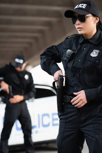 Policewoman dans des lunettes de soleil prendre pistolet de holster avec collègue et voiture sur fond flou — Photo de stock