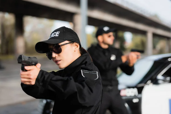 Policewoman en lunettes de soleil tenant pistolet près d'un collègue sur fond flou dans la rue urbaine — Photo de stock