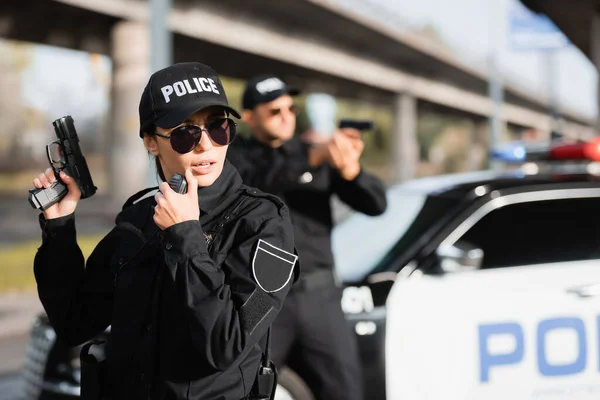 Poliziotta in occhiali da sole pistola in mano e utilizzando walkie talkie vicino collega e auto su sfondo sfocato — Foto stock