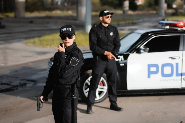 Poliziotta con pistola utilizzando walkie talkie vicino collega e auto della polizia su sfondo sfocato sulla strada urbana — Foto stock