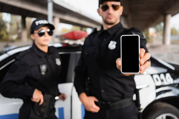 Smartphone avec écran blanc en main du policier près de collègue et voiture sur fond flou — Photo de stock