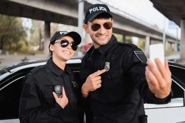 Cheerful police officers showing badge and walkie talkie while taking selfie on smartphone on blurred foreground outdoors — Stock Photo