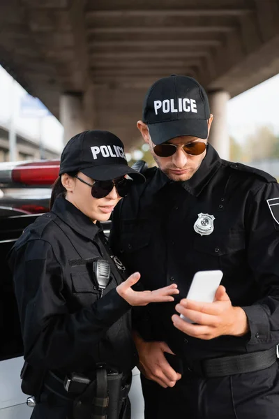 Mujer policía en gafas de sol apuntando a teléfono inteligente cerca de colega y coche sobre fondo borroso - foto de stock