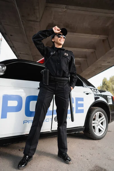 Весела поліцейська в сонцезахисних окулярах дивиться далеко біля поліцейського авто на відкритому повітрі — стокове фото