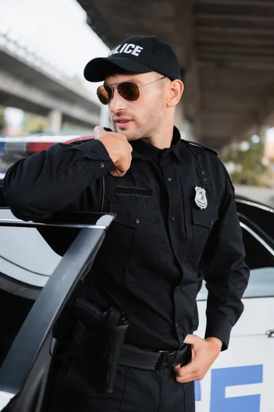 Policeman in sunglasses using walkie talkie near car on urban street — Stock Photo
