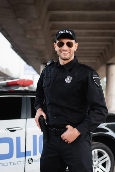 Усміхнений поліцейський у сонцезахисних окулярах тримає пістолет у кобурі біля машини на розмитому тлі — стокове фото