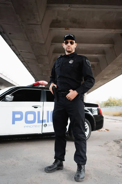 Officer of police holding gun near car on urban street — Stock Photo