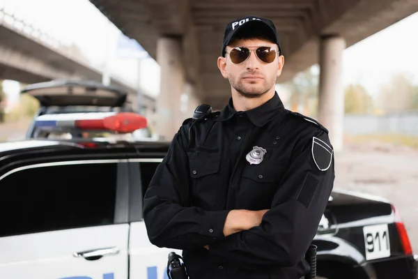 Police officer in sunglasses standing with crossed arms near auto on blurred background outdoors — Stock Photo