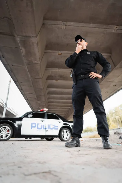 Low angle view of policeman in sunglasses standing with hand on hip near car on blurred background on urban street — Stock Photo