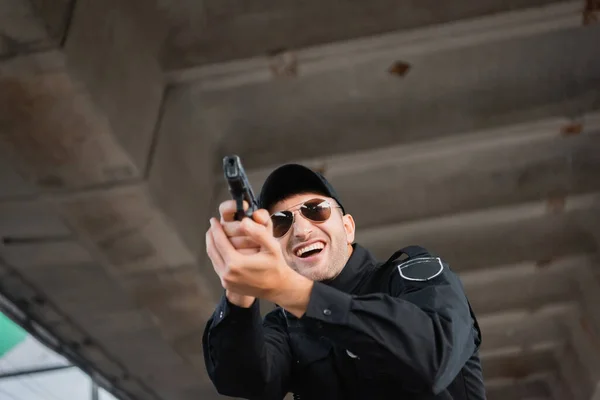 Low angle view of cheerful policeman in sunglasses holding gun on blurred foreground outdoors — Stock Photo