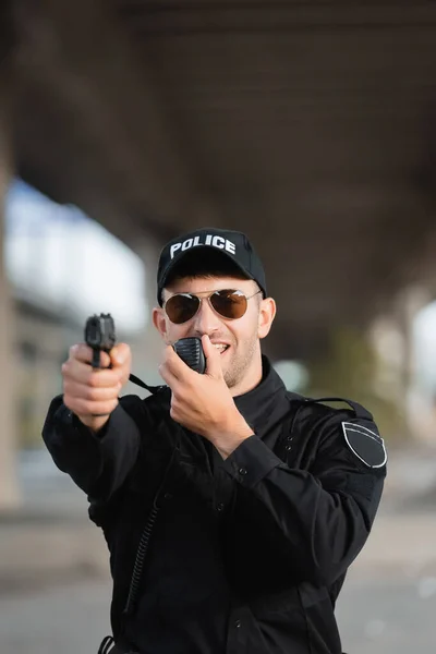 Police officer in sunglasses using walkie talkie and holding gun on blurred foreground outdoors — Stock Photo