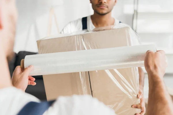 Partial view of worker wrapping box with stretch film near indian colleague — Stock Photo