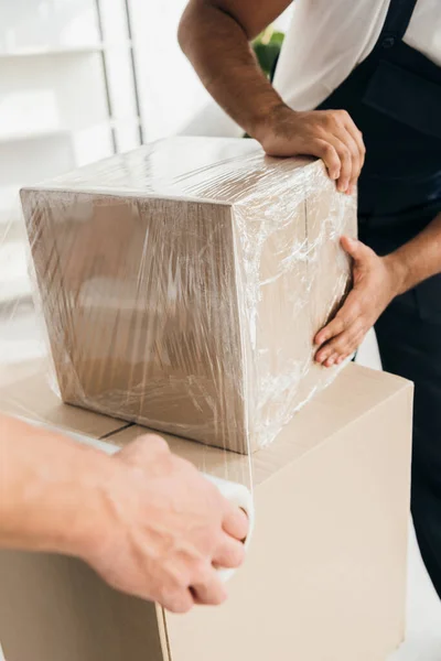 Partial view of mover holding wrapped in stretch film box near worker on blurred foreground — Stock Photo