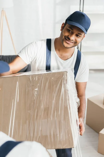 Happy indian mover holding wrapped in stretch film box near worker on blurred foreground — Stock Photo