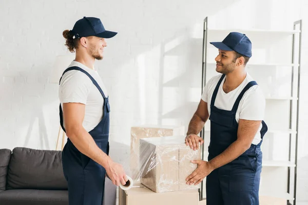 Side view of happy multicultural movers in overalls wrapping boxes with stretch film — Stock Photo