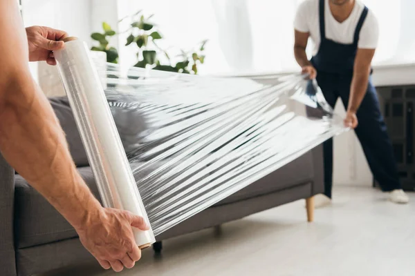 Cropped view of movers holding stretch film roll near sofa — Stock Photo