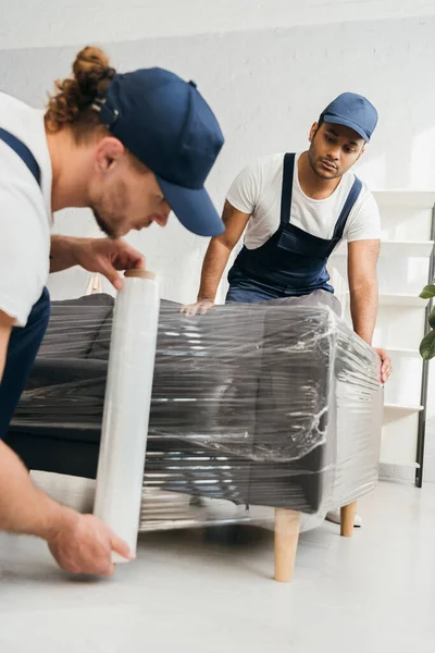 Indian mover looking at colleague holding stretch film roll near sofa on blurred foreground — Stock Photo