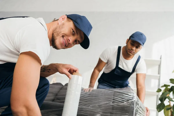 Cheerful and tattooed mover in cap holding stretch film roll near sofa and indian colleague on blurred background — Stock Photo