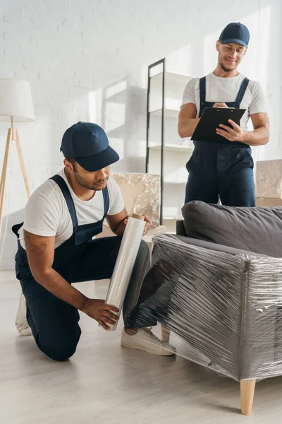 Indian mover in cap holding stretch film roll near sofa and worker with clipboard on blurred background — Stock Photo