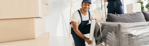 Fröhlicher indischer Mover in Mütze mit Stretchfolienrolle in der Nähe von Sofa und Arbeiter, Banner — Stockfoto