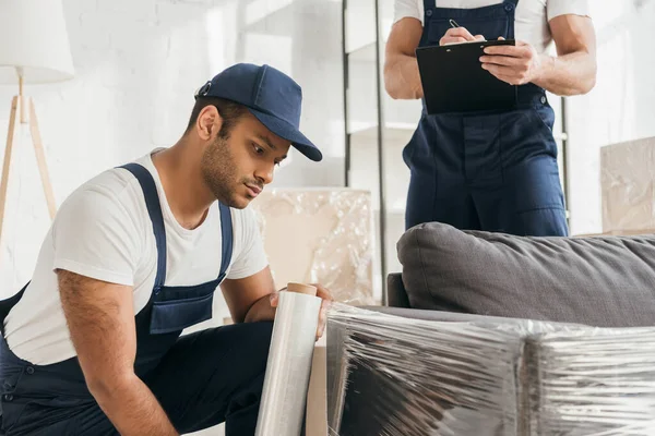 Traslochi indiani in cappuccio con pellicola estensibile rotolo vicino al divano e scrittura operaia sugli appunti — Foto stock