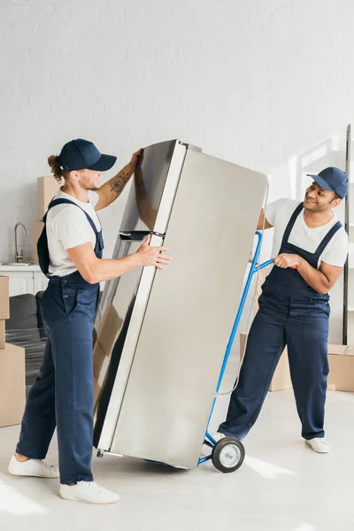 Cheerful indian mover looking at coworker while moving fridge in apartment — Stock Photo