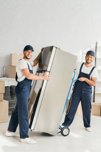 Cheerful multicultural movers standing near fridge in apartment — Stock Photo