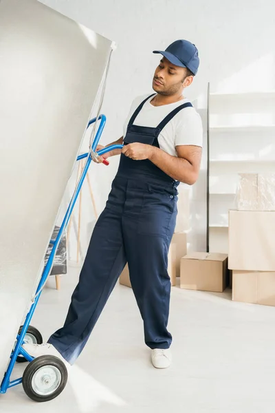 Strong indian workman holding hand truck while moving fridge in apartment — Stock Photo