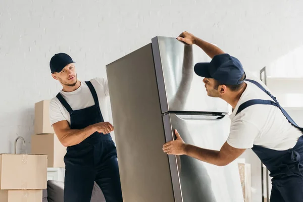 Multicultural movers in uniform moving fridge in apartment — Stock Photo